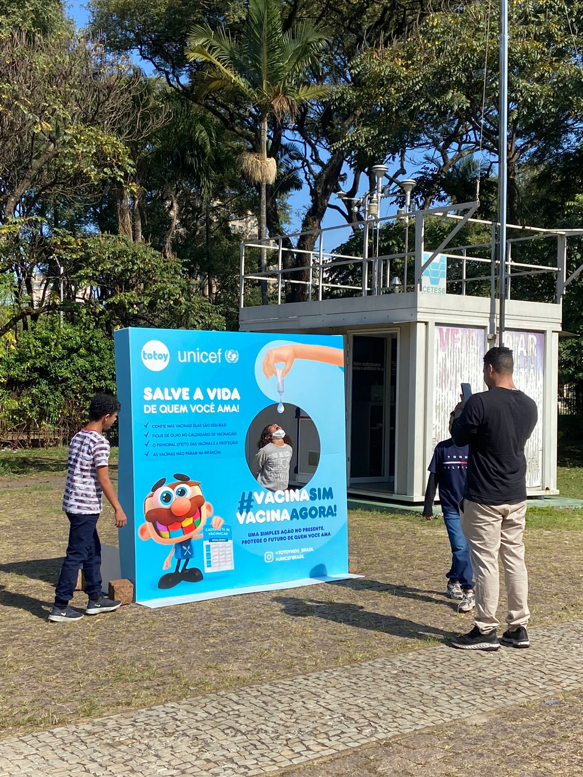 Imagem com uma família tirando foto no Backdrop da campanha "Vacina Sim! Vacina Agora!"